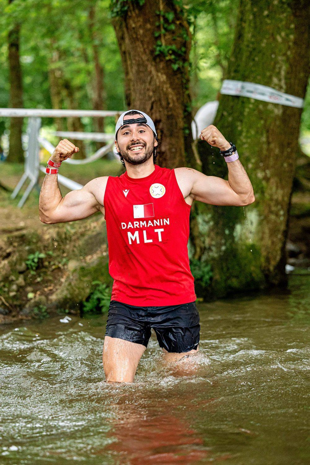 Isaac Darmanin in a red Malta race vest, flexing while wading through a river during a race